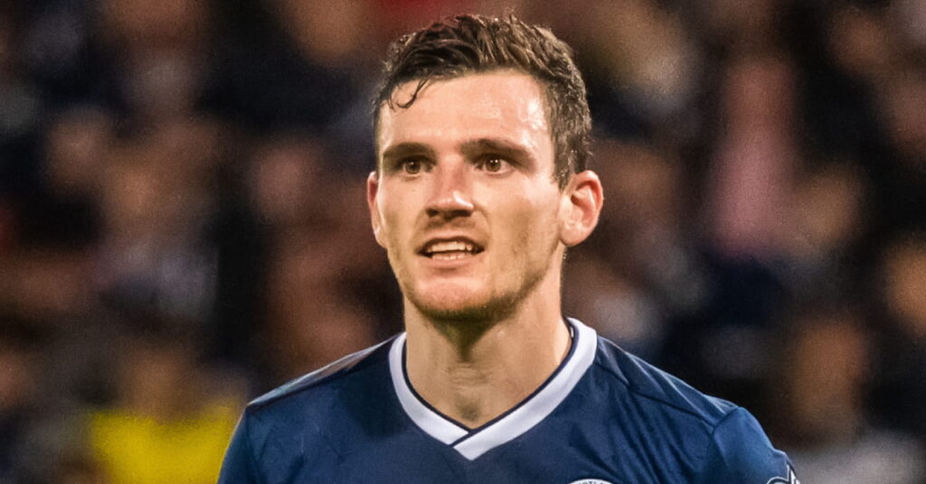 O jogador de futebol Andy Robertson joga em campo vestindo uma camisa de futebol da seleção escocesa com gola branca e um top azul escuro com a bandeira nacional em amarelo no peito. Robertson olha para a direita