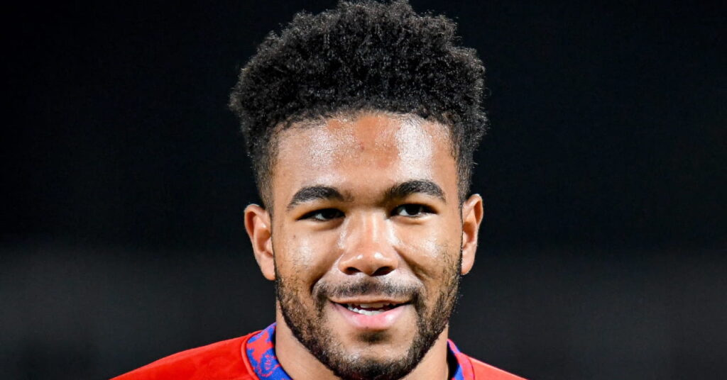 O jogador de futebol Reece James sorri no campo do estádio. Este jogador de futebol está vestindo uma camisa de treino da seleção inglesa de cor azul vermelha