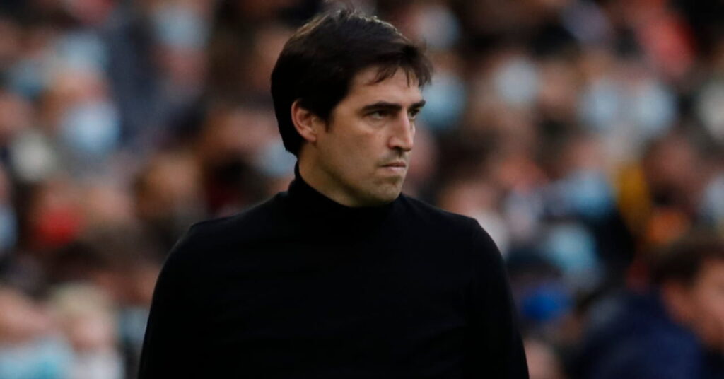 Football manager Andoni Iraola looking to his left with a crowd blurrily visible behind him. The coach is wearing a black jumper