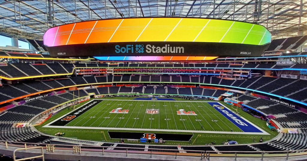Foto da Copa do Mundo de 2026 e do estádio de futebol americano Sophie Stadium em Los Angeles, mostrando o interior do estádio com cores vivas, fileiras de assentos pretos em concreto e uma grande tela de vídeo circular mostrando o campo listrado verde sob um céu azul