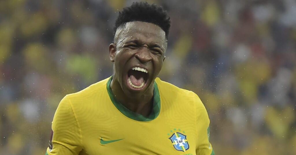 A photo of footballer Vinicius Junior shouting in celebration and smiling while wearing a Brazil national football team shirt in a stadium. The shirt is yellow with green trim and has a green Nike tick and Brazil badge on the chest