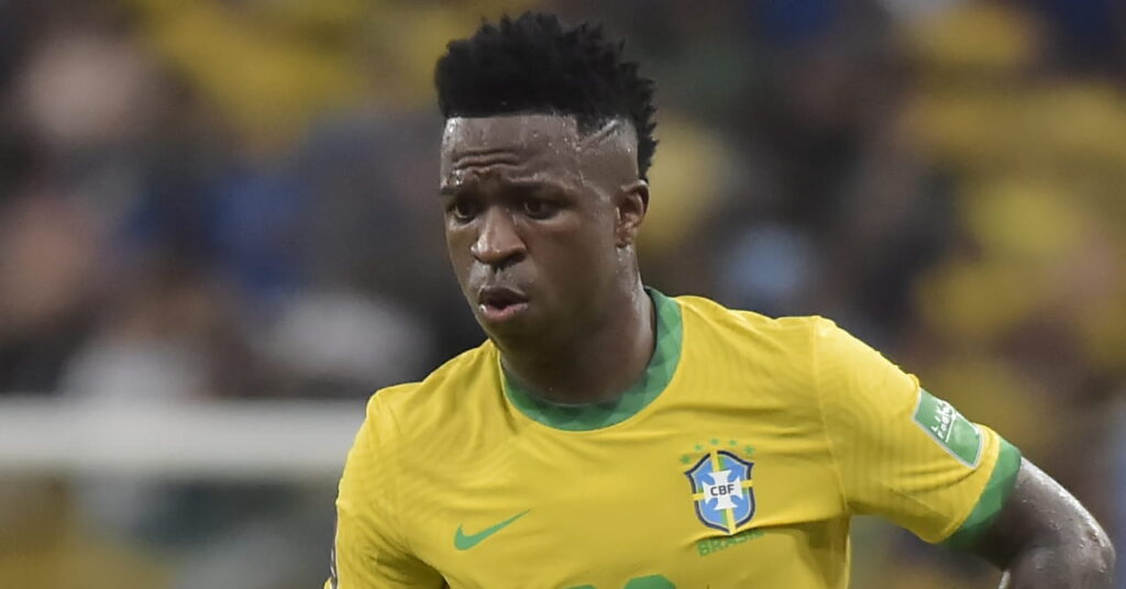 A photo of footballer Vinicius Junior looking downwards while wearing a Brazil national football team shirt in a stadium. The shirt is yellow with green trim and has a green Nike tick and Brazil badge on the chest