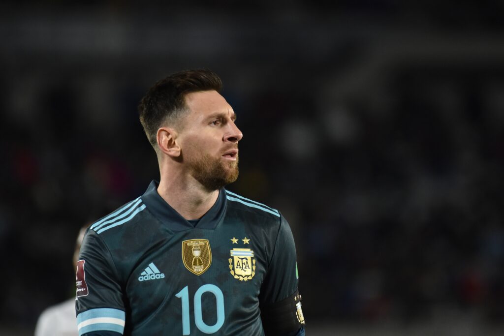 Uma foto do jogador de futebol Lionel Messi olhando para a esquerda em um campo. Messi veste uma camisa azul escura da seleção argentina de futebol com uma faixa azul clara no ombro em um estádio e o número 10 da Adidas em azul claro no peito ao lado de um distintivo dourado da seleção nacional.