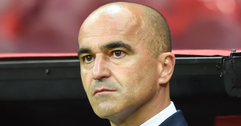 A photo of Portugal manager Roberto Martinez looking to his right while standing in a technical area constructed of black metal on a football pitch. The head coach is wearing a dark suit jacket and white shirt