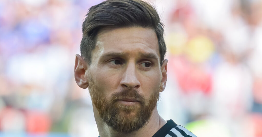 A photo of footballer Lionel Messi looking to his left on a pitch. Messi is wearing an Argentina national football team shirt in dark blue with light blue stripes down the shoulders in a stadium setting with his number 10 and the Adidas in light blue on the chest next to the gold national team badge