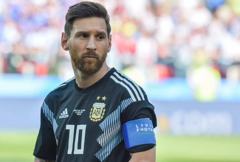 A photo of footballer Lionel Messi looking to his left on a pitch. Messi is wearing an Argentina national football team shirt in dark blue with light blue stripes down the shoulders in a stadium setting with his number 10 and the Adidas in light blue on the chest next to the gold national team badge