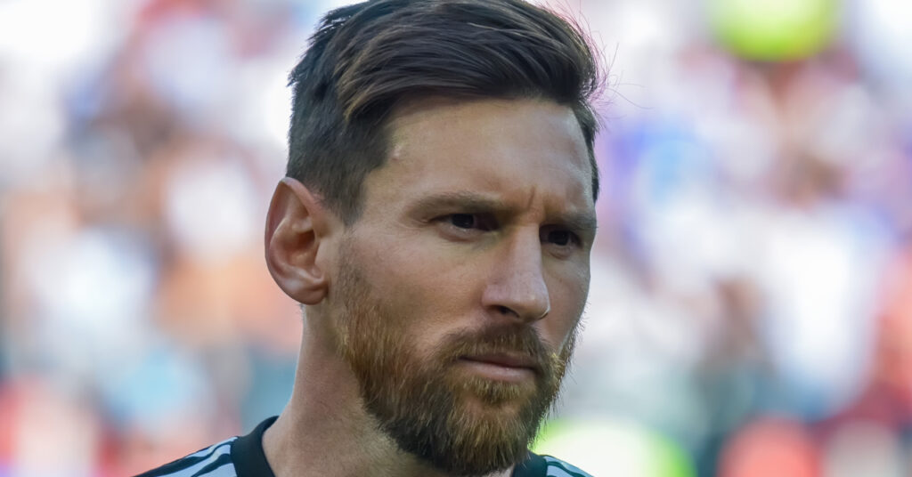 A photo of footballer Lionel Messi looking to his left on a pitch. Messi is wearing an Argentina national football team shirt in white with blue stripes down the centre with a gold national badge on the chest and a blue captain armband and black trim