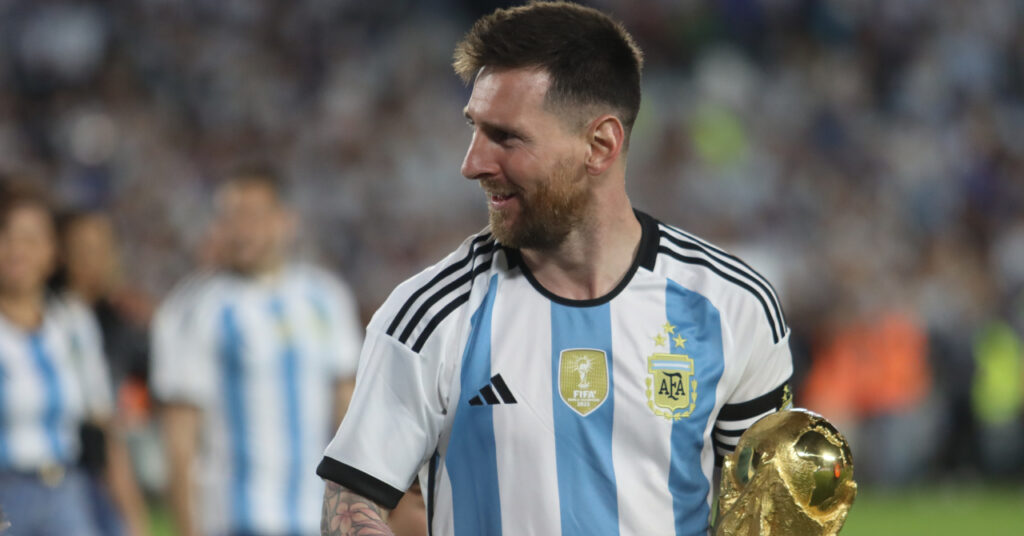 O jogador de futebol Lionel Messi sorrindo em campo, olhando para a direita e segurando a mão direita para acenar. Messi veste uma camisa da seleção argentina de futebol branca com listras azuis com um emblema nacional dourado no peito e segura o troféu da Copa do Mundo à sua esquerda com um campo e uma multidão vagamente visíveis ao fundo.