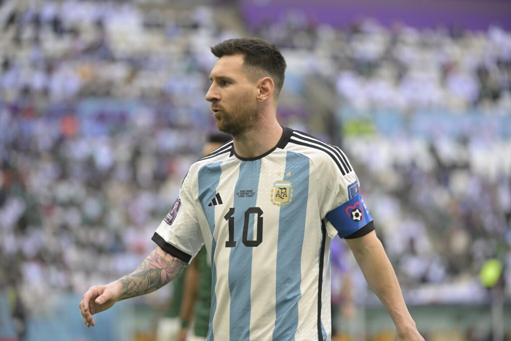 A photo of footballer Lionel Messi looking to his right on a pitch. Messi is wearing an Argentina national football team shirt in white with blue stripes down the centre with a gold national badge on the chest and a b;ue captain armband and black trim
