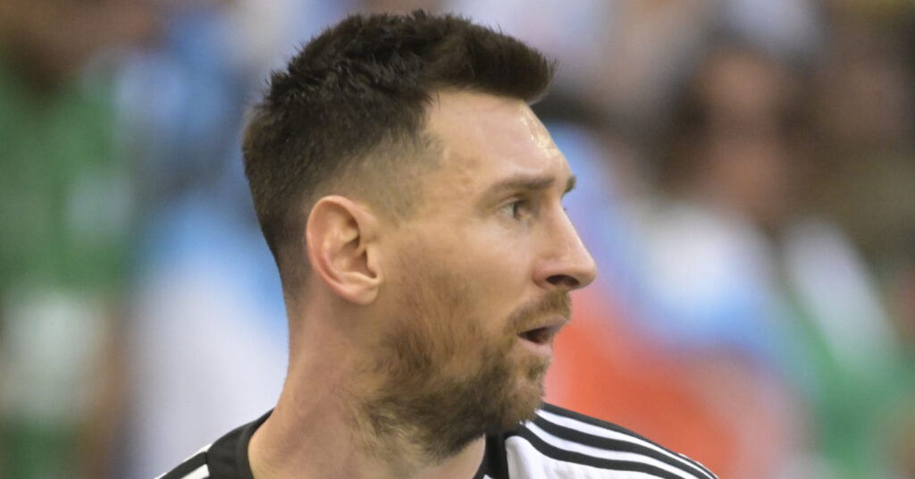 A photo of footballer Lionel Messi looking to his left on a pitch. Messi is wearing an Argentina national football team shirt in white with blue stripes down the centre with a gold national badge on the chest and a blue captain armband and black trim