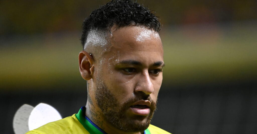 Uma foto de Neymar, um jogador de futebol vestindo uma camisa da seleção brasileira de futebol, olhando para um estádio. A camisa é amarela com detalhes verdes e traz marca Nike verde e escudo do Brasil no peito.