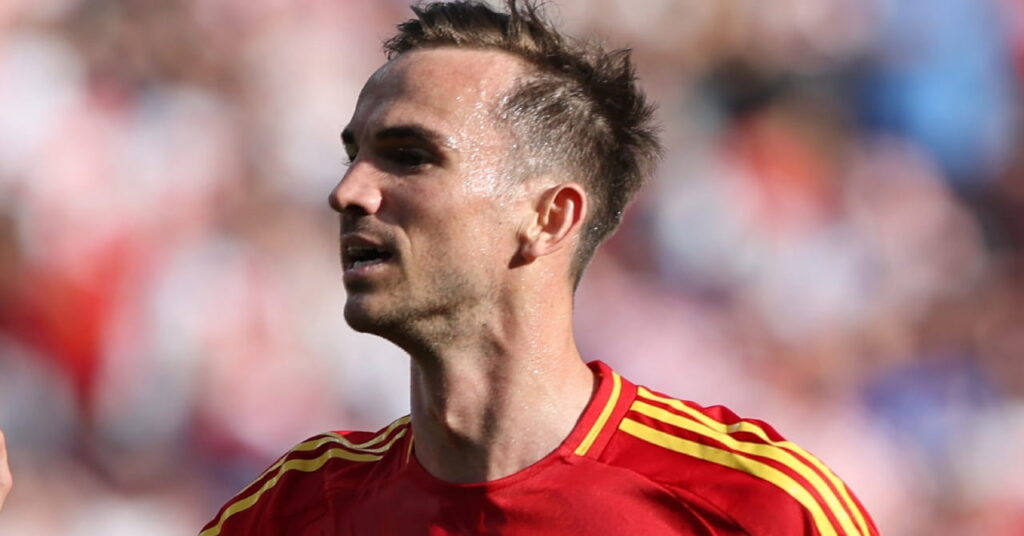A photo of footballer Fabian Ruiz looking to his right while standing on a pitch playing for the Spain national football team in a red shirt with yellow lines on the shoulders