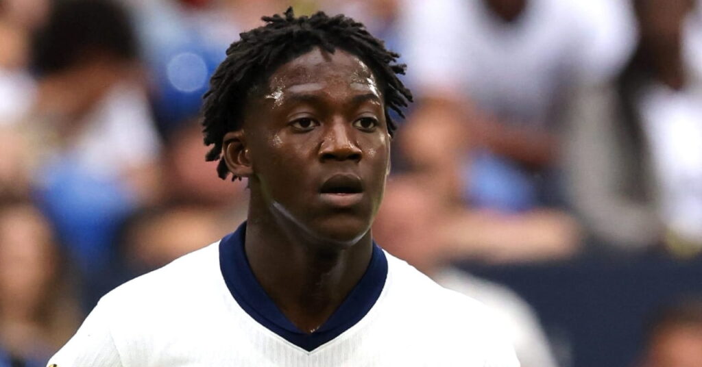 Footballer Kobbie Mainoo pulling an open-mouthed look of concentration on a stadium pitch. The football player is wearing the England national team shirt of white with blue collar