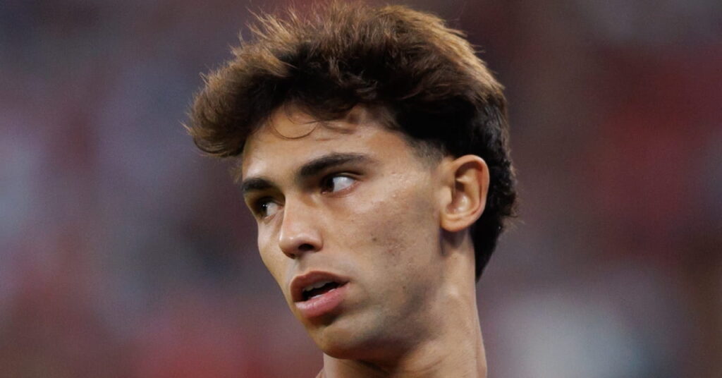 A photo of footballer Joao Felix looking to his right while on a football pitch. The football player is wearing a Portugal national football team kit in red with green and black trim and a white nike logo and number on his chest in a stadium setting