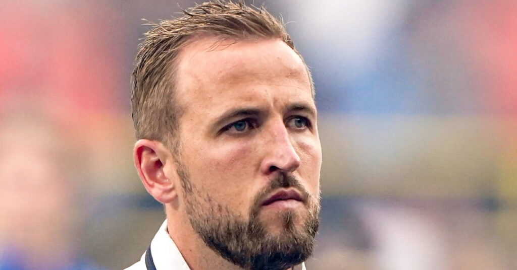 Footballer Harry Kane looking to his left on a stadium pitch. The football player is wearing the England national team training top of white with blue collar