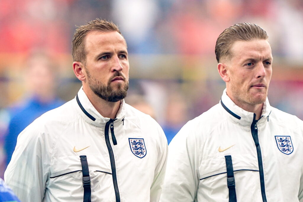 O jogador de futebol olha para a esquerda no campo do Harry Kane Stadium. Este jogador de futebol está vestindo um top de treino da seleção inglesa com cor branca e gola azul