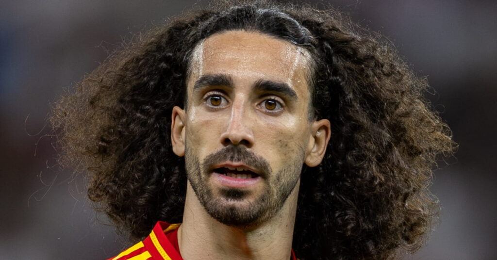 A photo of footballer Marc Cucurella looking to his right while standing on a pitch wearing the Spain national team football shirt of red with yellow trim. The football player's number 24 is on his chest in yellow along with the Adidas logo and national team badge. A crowd is blurrily visible in the background