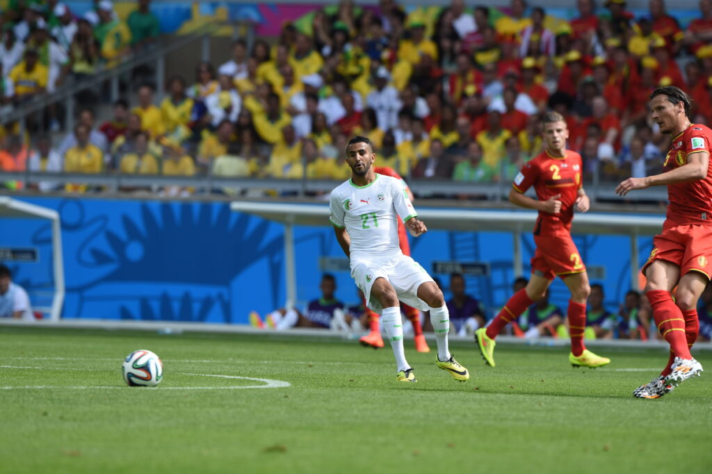 Uma foto do jogador de futebol Riyad Mahrez chutando uma bola de futebol enquanto jogava pela seleção argelina de futebol em um campo gramado. Mahrez usa um kit branco com detalhes verdes e chuteiras Nike amarelas e pretas, com um logotipo vermelho da Puma no peito e emblemas da Argélia e do futebol em ambos os lados, ao lado de seu número 21 em verde. Uma infinidade de cores amarelas e opções em uma área técnica são levemente visíveis no fundo contra um fundo azul. Os dois jogadores com o uniforme belga são visíveis à esquerda de Mahrez, principalmente vermelhos com detalhes e cores amarelas