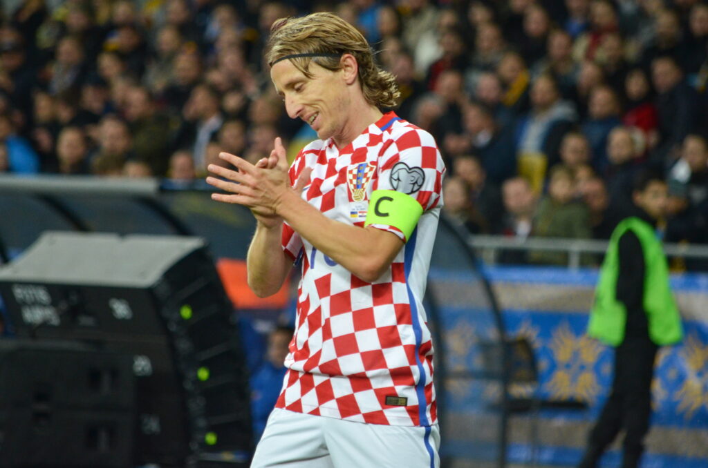 O jogador de futebol Luka Modric olha para baixo e bate palmas enquanto joga dentro do estádio com a camisa vermelha e branca da seleção croata de futebol