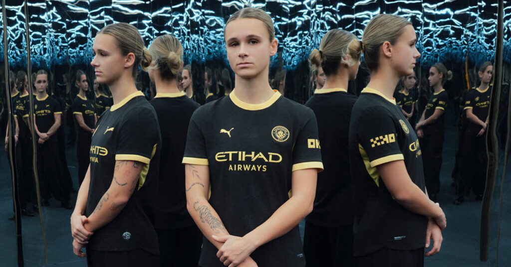 Man City footballer Kerstin Casparij modelling the predominantly black away kit of Manchester City Football Club with a yellow trim and Etihad Airways on the front in a darkened room