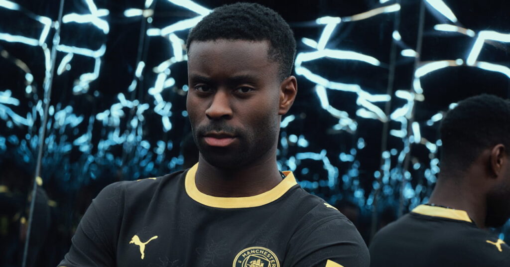 Man City footballer Marc Guehi modelling the predominantly black away kit of Manchester City Football Club with a yellow trim and Etihad Airways on the front in a darkened room