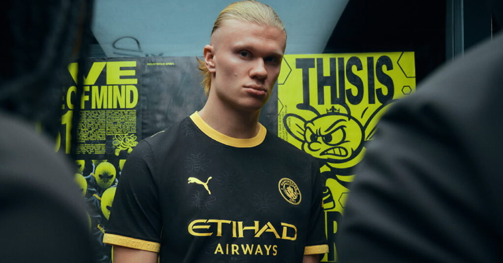Man City footballer Erling Haaland modelling the predominantly black away kit of Manchester City Football Club with a yellow trim and Etihad Airways on the front in a darkened room