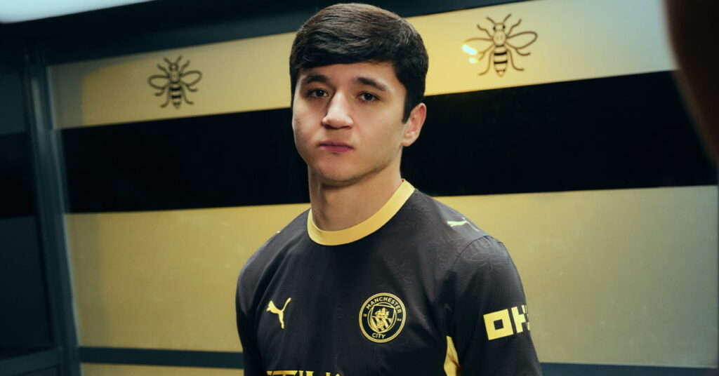 Footballer Abdukodir Khusanov modelling the predominantly black away kit of Manchester City Football Club with a yellow trim and Etihad Airways on the front in a darkened room