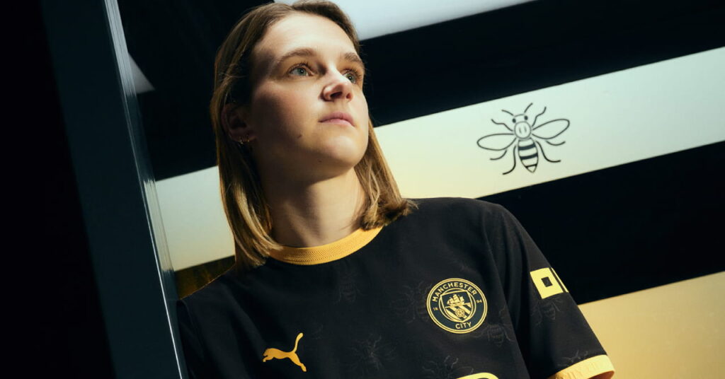 Man City footballer Vivianne Miedema modelling the predominantly black away kit of Manchester City Football Club with a yellow trim and Etihad Airways on the front in a darkened room