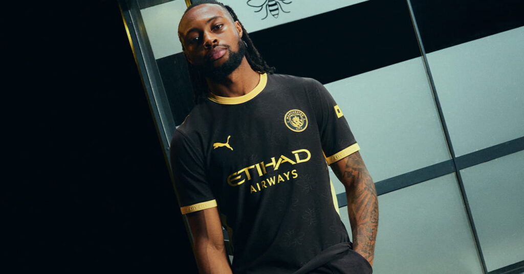 Manchester City footballer Antoine Semenyo modelling the predominantly black away kit of Man City Football Club with a yellow trim and Etihad Airways on the front in a darkened room