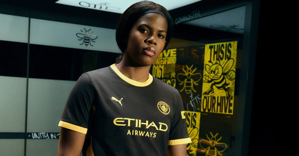 Man City footballer Bunny Shaw modelling the predominantly black away kit of Manchester City Football Club with a yellow trim and Etihad Airways on the front in a darkened room