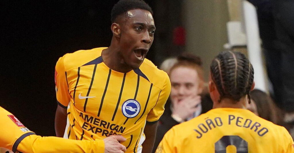 Footballer Danny Welbeck jumping and shouting to celebrate a goal while playing for Brighton and Hove Albion Football Club in a yellow kit with dark blue stripes and teammates congratulating him