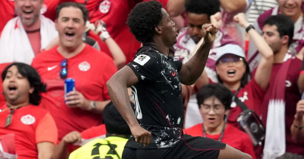 O futebol Jonathan David, vestindo um uniforme predominantemente preto com detalhes em branco e vermelho, pula no ar enquanto vai para a direita para comemorar um gol durante um jogo pela seleção canadense de futebol na Copa do Mundo de 2026 diante de torcedores entusiasmados