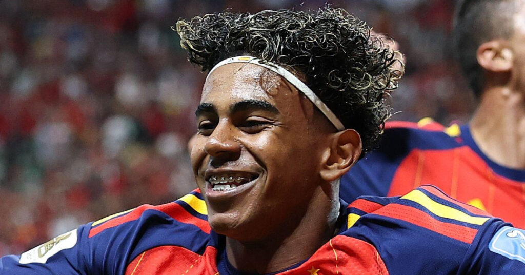 Footballer Lamine Yamal smiling while playing in a stadium for the Spain national football team, wearing their home colours of red, yellow and blue