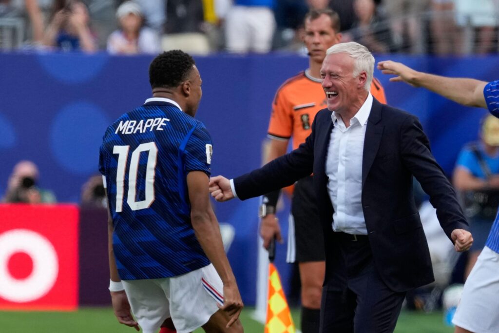 Footballer Kylian Mbappe looking to his right and celebrating on a pitch while wearing a blue France national football team shirt with his name and number 10 on the back at the World Cup 2026, celebrating with head coach Didier Deschamps, who is smiling and wearing a dark suit jacket and white shirt