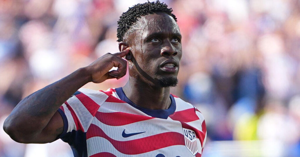 O jogador de futebol Florin Balogun leva a mão esquerda à orelha esquerda enquanto comemora um gol durante a Copa do Mundo de 2026 com o uniforme da seleção nacional de futebol da USMNT com detalhes azuis nas listras vermelhas e brancas.