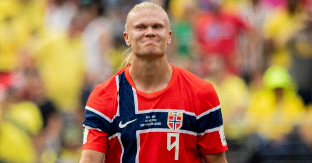Footballer Erling Haaland smiling on a pitch during the World Cup 2026 while wearing the Norway national team football kit of a red shirt with blue and white cross and his number in white on the shirt with a crowd blurrily visible in the background
