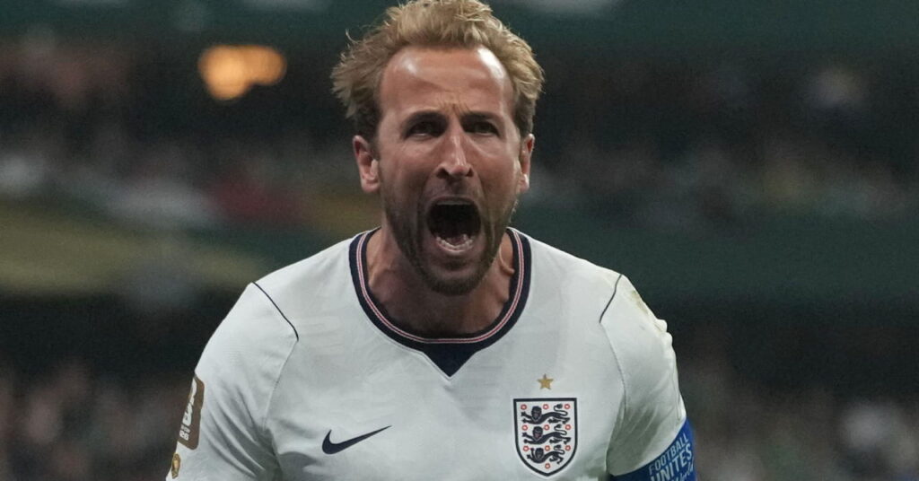 O jogador de futebol Harry Kane ruge e corre enquanto comemora um gol durante a Copa do Mundo de 2026, enquanto o kit de futebol da seleção inglesa consiste em uma camisa branca com detalhes em azul e seu número em uma camisa vermelha com shorts azul escuro e o emblema da Inglaterra com detalhes em branco e seu número em branco.