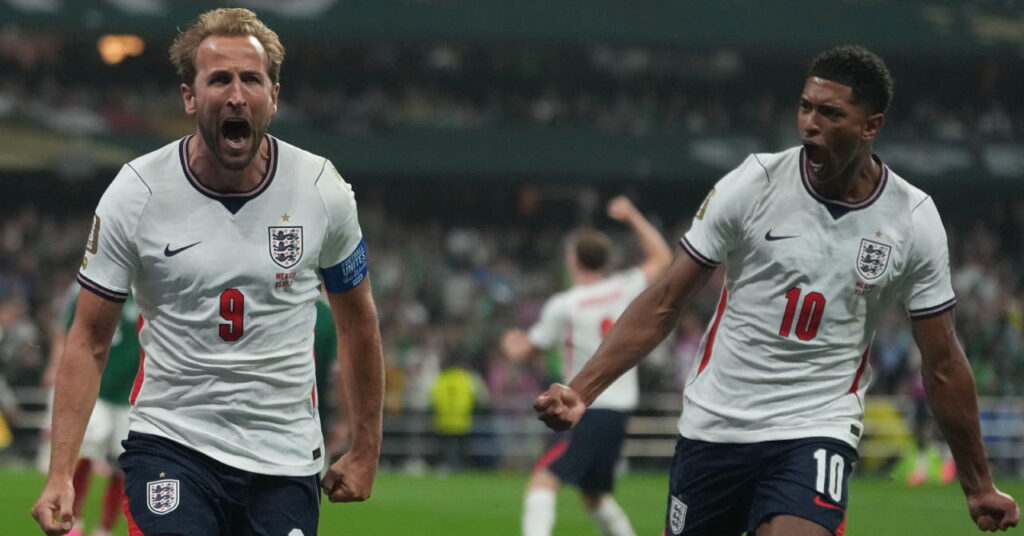 Footballers Harry Kane and Jude Bellingham roaring and running while celebrating a goal during the World Cup 2026 while wearing the England national team football kit of a white shirt with blue trim and their numbers in red on the shirt with dark blue shorts and white trim featuring the England badge and their numbers in white