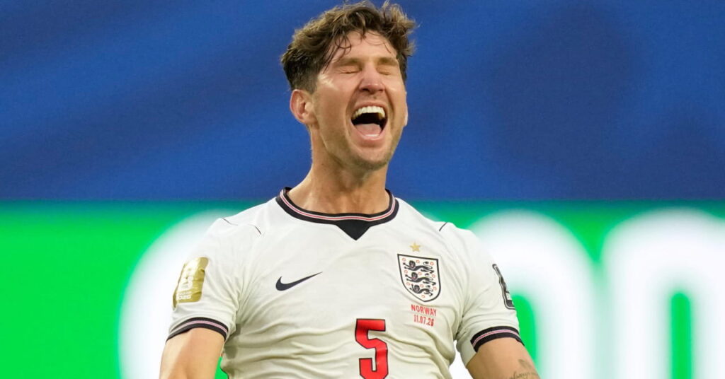 Footballer John Stones kneeling on a pitch with his arms outstretched smiling and shouting in celebration while playing in the England national football team kit of white with blue and red details at the World Cup 2026