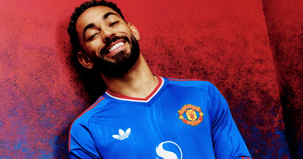 Footballer Matheus Cunha posing in the Manchester United Football Club blue away shirt with red and white trim and the club badge and Adidas logo on the chest against a red background