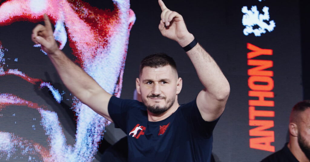 O boxeador peso pesado Cristian Prenga levanta os braços com uma camisa preta no palco como parte da pesagem para sua luta de boxe peso pesado com Anthony Joshua
