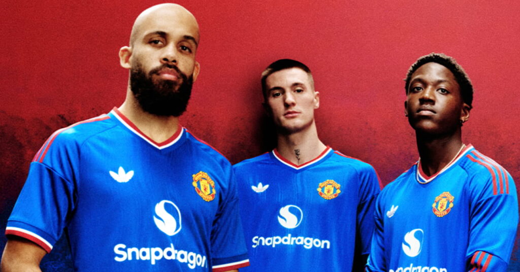Footballers Bryan Mbeumo, Benjamin Sesko and Kobbie Mainoo posing in the Manchester United Football Club blue away shirt with red and white trim and the club badge and Adidas logo on the chest against a red background
