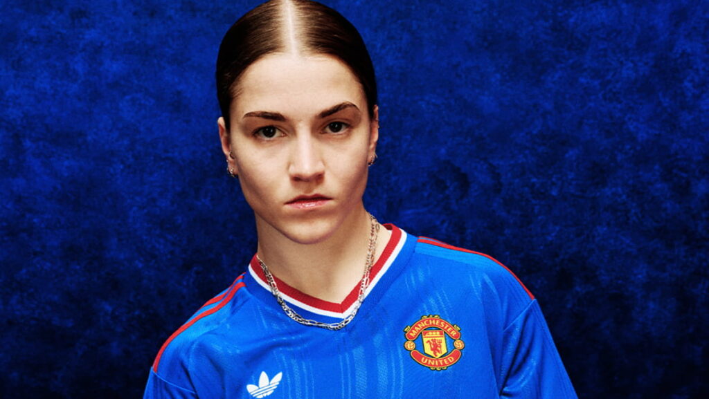 A jogadora de futebol Jess Park posa no Manchester United Football Club vestindo uma camisa azul com detalhes em vermelho e branco e o escudo do clube e o logotipo da adidas no peito contra um fundo azul