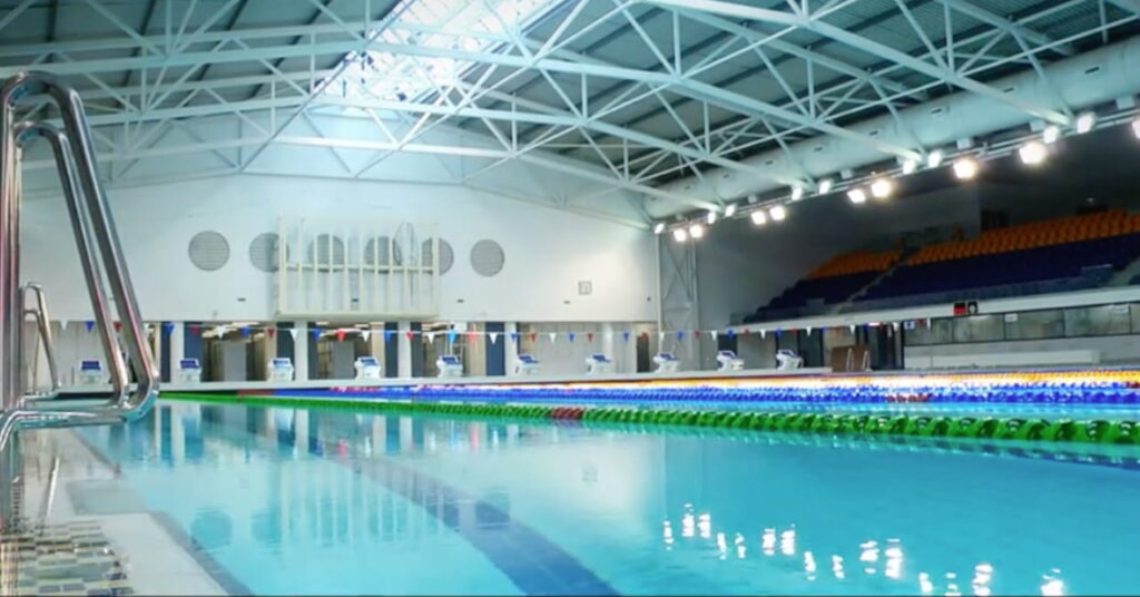 O local da Tollcross International Swimming Pool em Glasgow para os Jogos da Commonwealth de 2026 mostrando perto da água e assentos para espectadores
