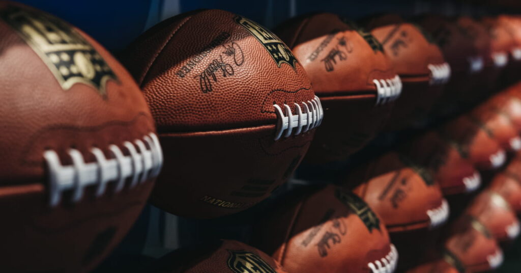 A close-up of brown footballs used in NFL American football, set on rows in a darkened room with white stitching and the competition logo in black and gold