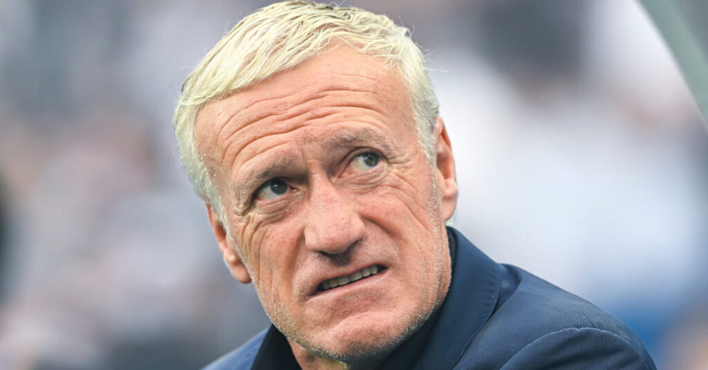 O técnico de futebol Didier Deschamps olha para a esquerda dentro de um estádio de futebol enquanto o técnico da seleção francesa veste um paletó escuro e camisa combinando.