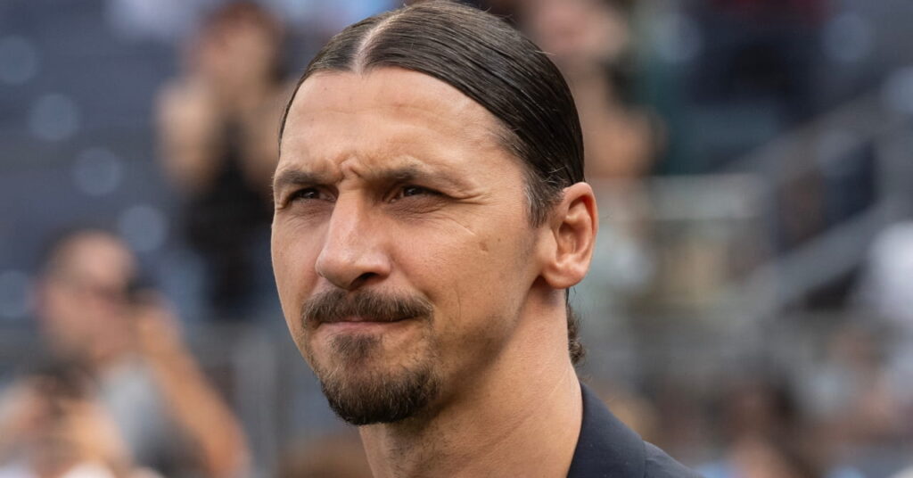 Former footballer Zlatan Ibrahimovic looking sternly to his right in a football stadium while wearing a dark suit jacket and top with a crowd blurrily visible in the background