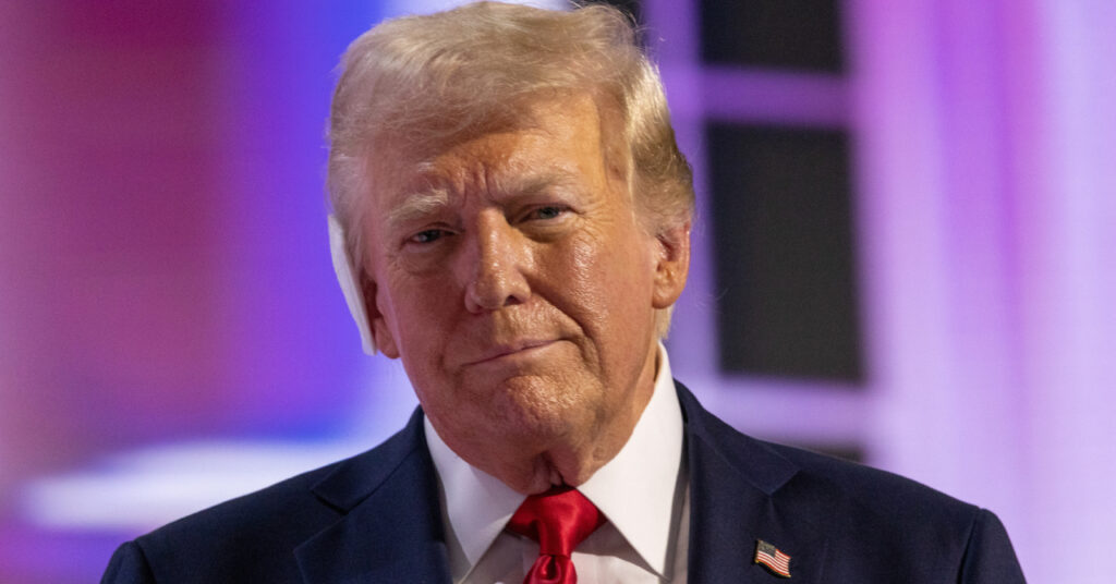 United States president Donald Trump standing up in a suit comprising a dark blue jacket, red tie and white shirt with the USA flag on a pin badge on the chest of the president