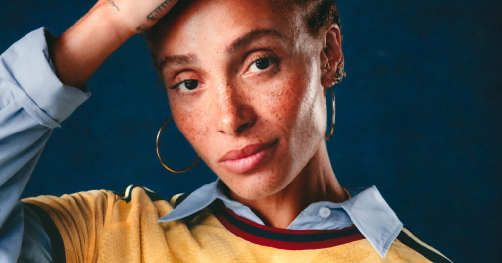 Model and actress Adwoa Aboah posing in the third shirt of Premier League team Arsenal Football Club while accompanying the yellow shirt with a blue collared formal shirt and raising her right hand to her head