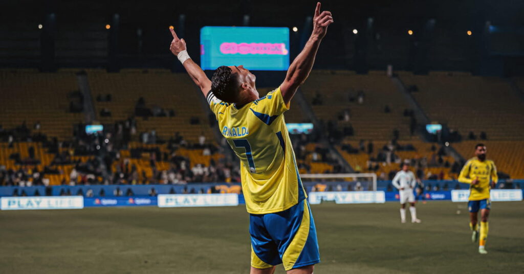 Portugal footballer Cristiano Ronaldo raising both arms in celebration in a stadium while playing for Saudi Pro League team Al Nassr Football Club in a yellow and blue kit with his surname and number seven on the back